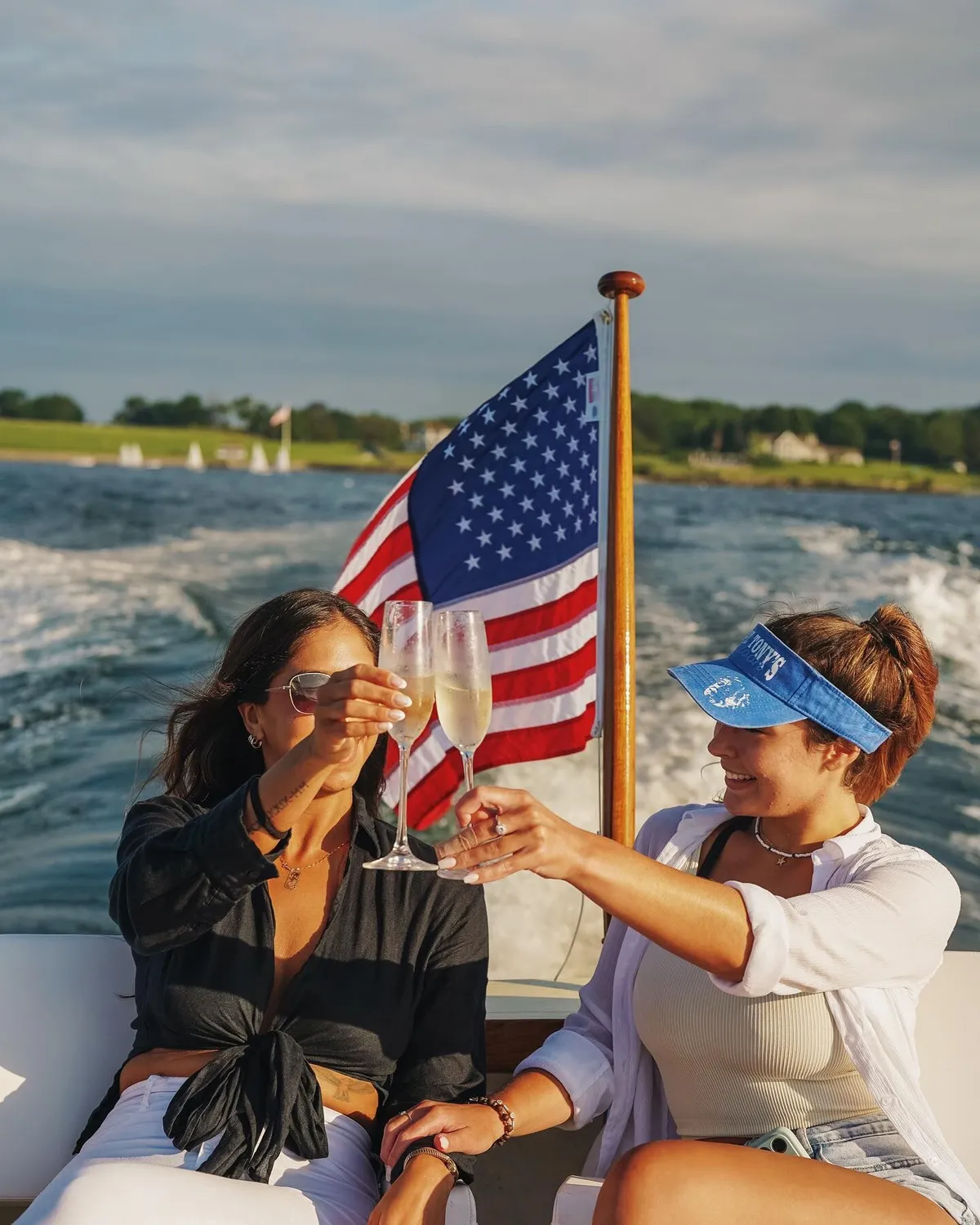 Two women clinking champagne glasses on a moving boat with an American flag and water behind them.