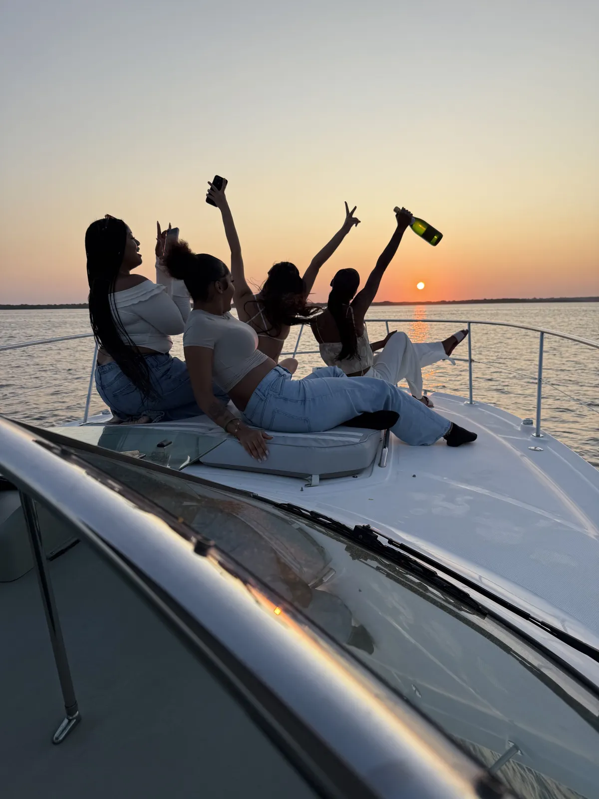Four women cheer on a boat bow at sunset while one raises a green champagne bottle.