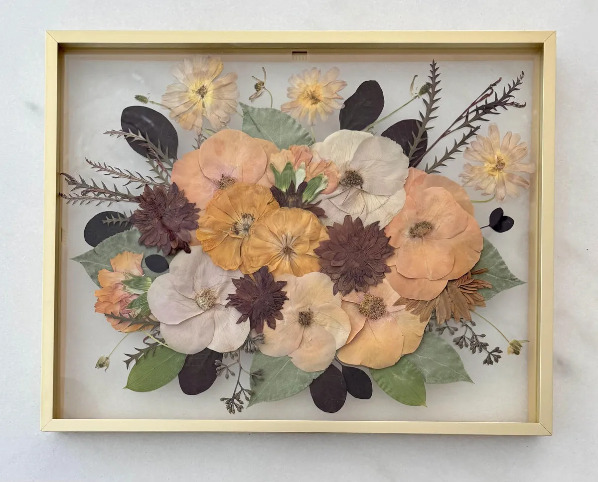 Pressed dried flowers in thin gold frame displayed flat on white marble surface.