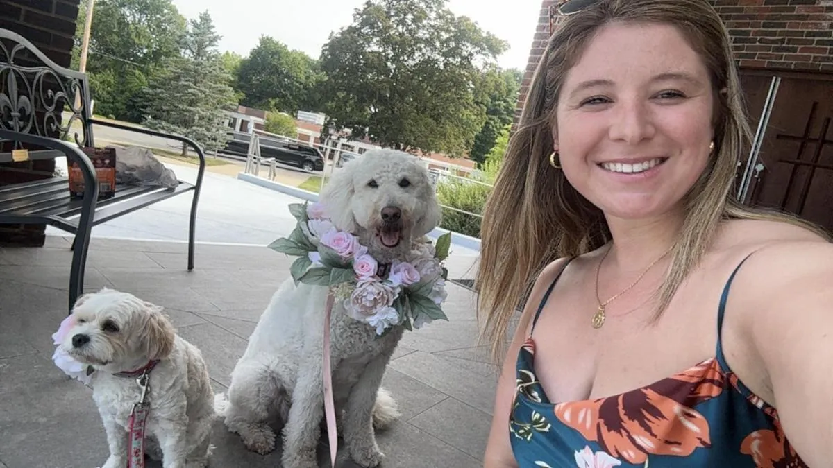 Smiling woman in a floral dress takes a selfie with two white dogs wearing pink floral collars on a patio.
